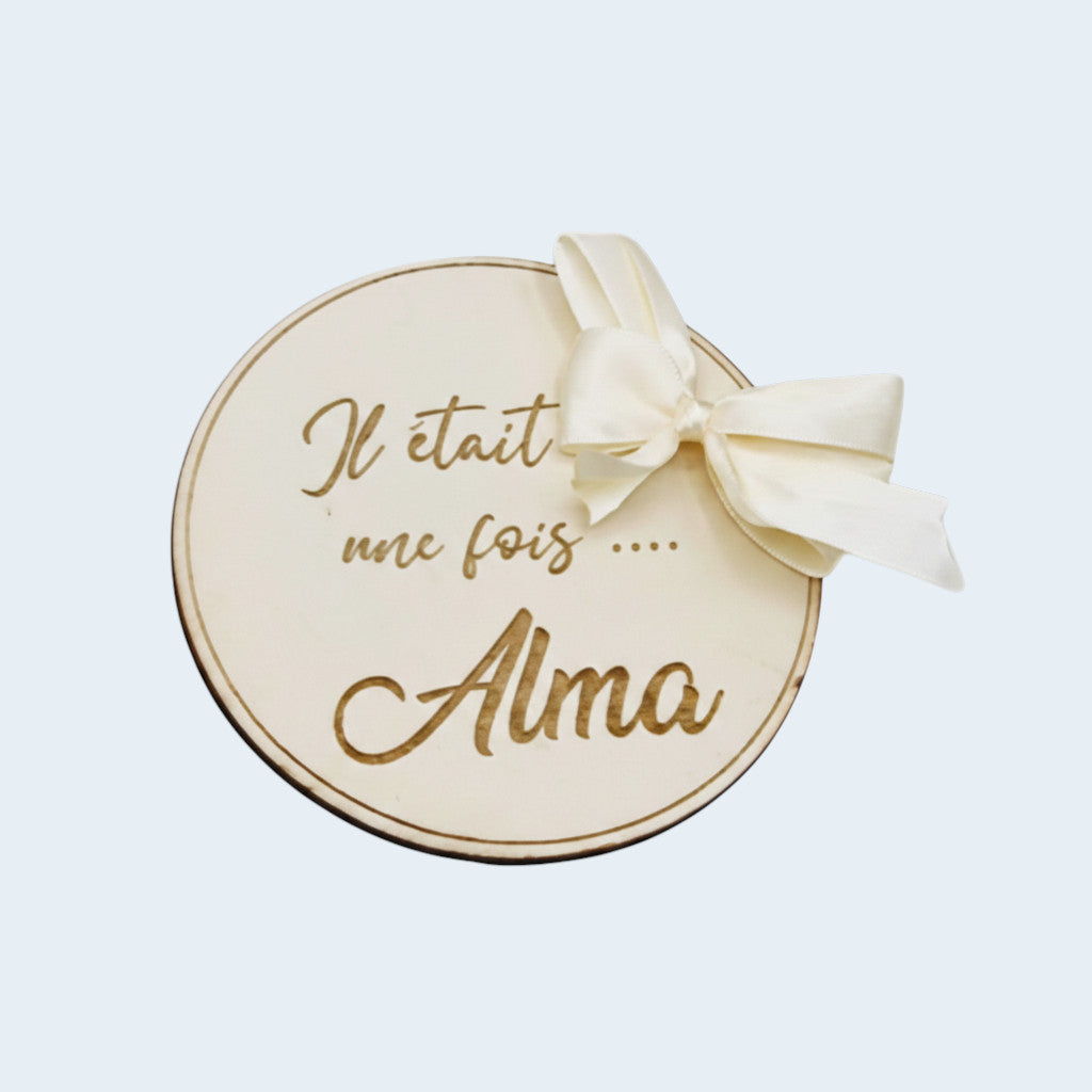 Carte de présentation naissance ronde en bois gravé avec prénom personnalisé et grand nœud en satin crème, style bohème chic.